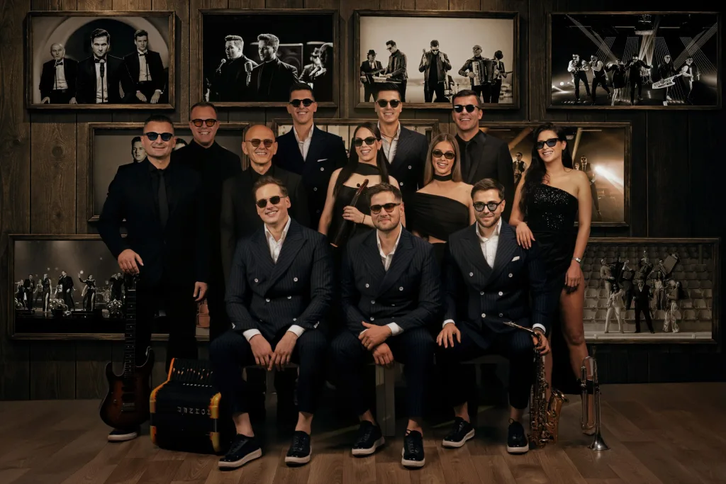 CHEGI BEND — članovi benda sa gitarom, harmonikom i saksofonom, ispred zida sa uramljenim fotografijama
